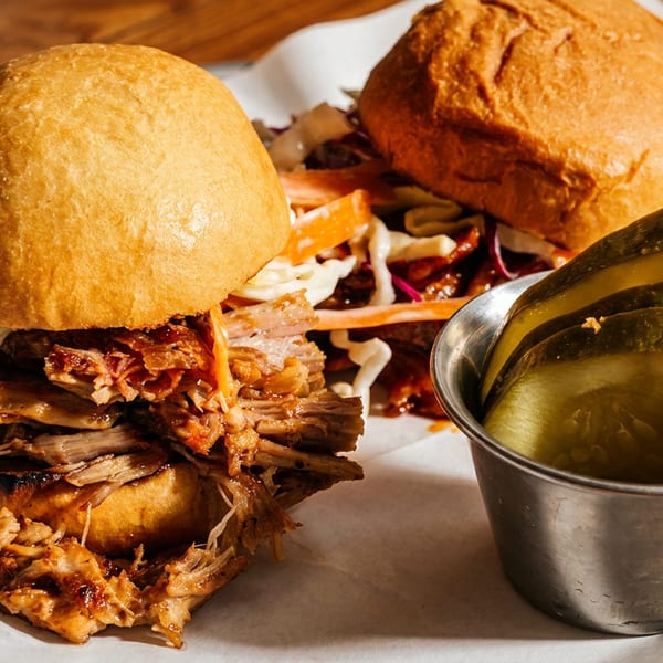 Pulled pork sandwich with coleslaw and pickles in a metal cup on white paper.