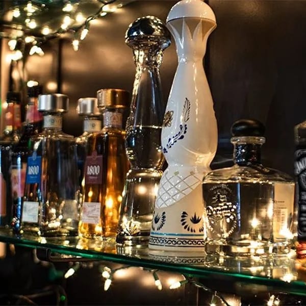 Assorted liquor bottles, including tequila and whiskey, on a glass shelf with string lights.