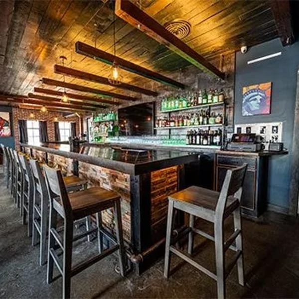 Rustic bar interior with wooden ceiling, brick-faced bar, and rows of bar stools.