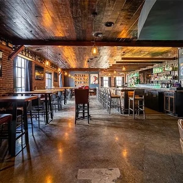 Spacious bar interior with brick walls, wooden ceilings, high-top tables, and a long bar with stools.