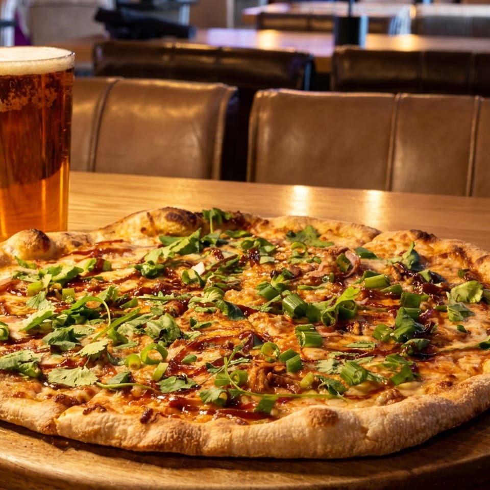 A BBQ chicken pizza with cilantro and green onions, next to a pint of beer on a wooden table.