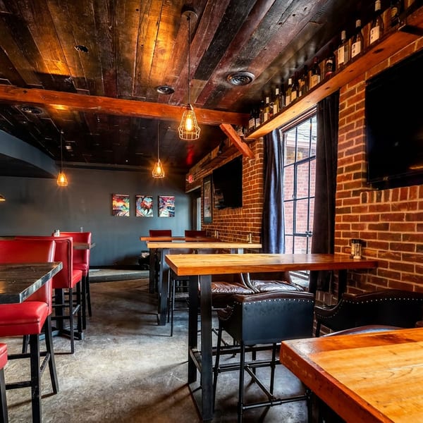 A dimly lit restaurant interior with red chairs, wooden tables, brick walls, and a dark wood ceiling.