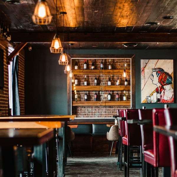 Warmly lit restaurant interior with brick walls, wooden ceiling, wine display, and red bar stools.