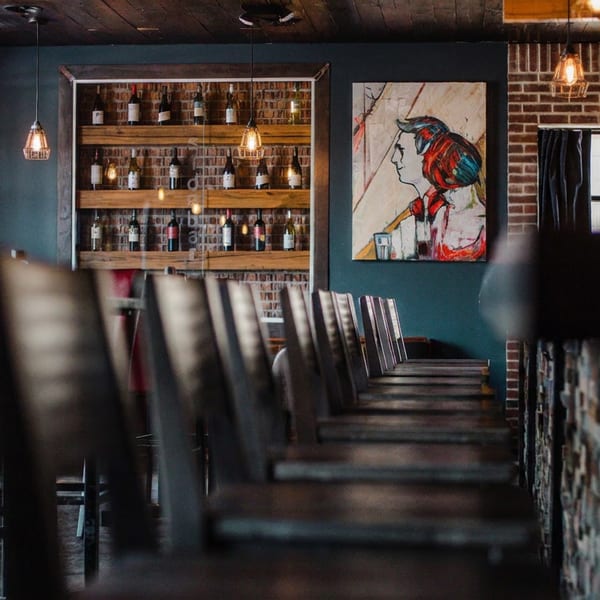 Restaurant interior with brick walls, wine bottles on shelves, modern art, and empty bar stools.