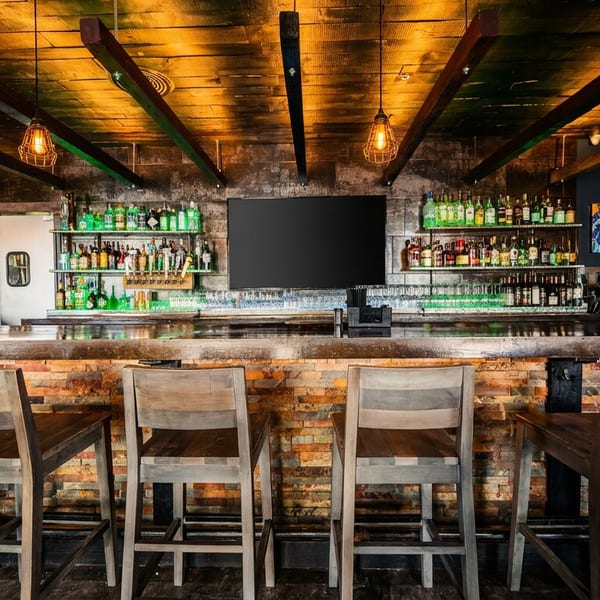 A rustic bar with wooden stools, shelves of liquor bottles, and a large TV screen.