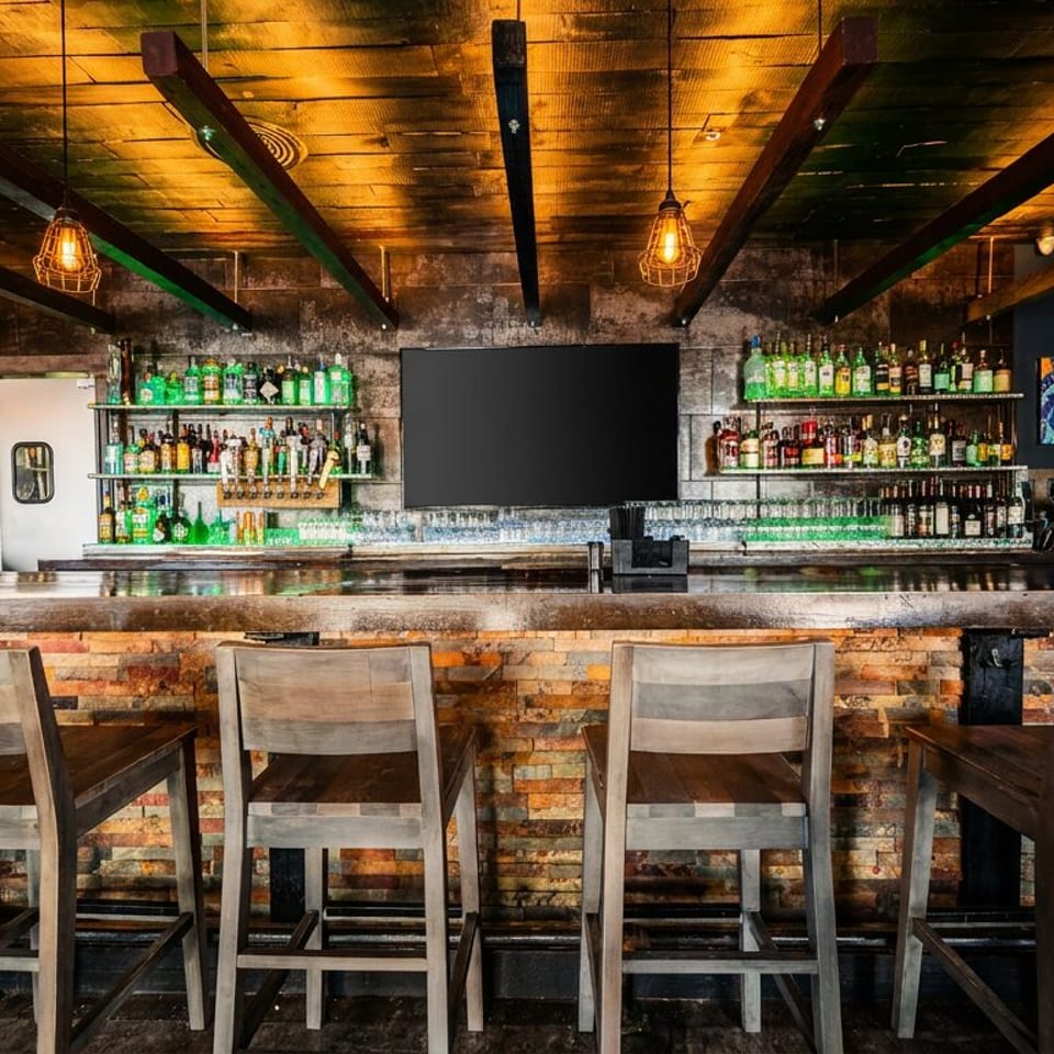 A rustic bar with wooden stools, a fully stocked liquor shelf, and a large TV screen.