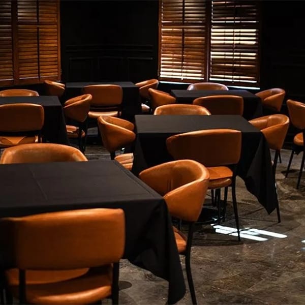 Empty restaurant dining room with black-covered tables and brown leather chairs, illuminated by window light.