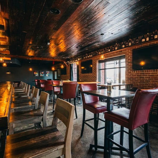 Warmly lit bar interior with a long wooden bar, high-top tables, and brick walls.