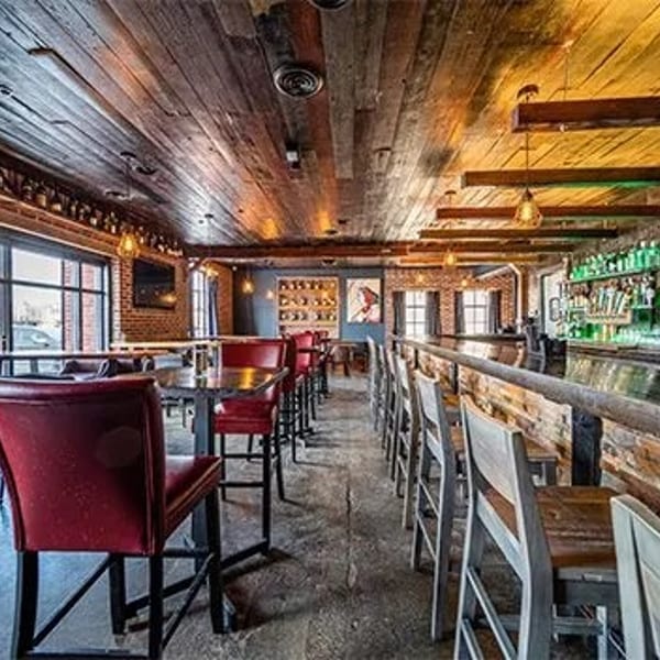 Rustic bar interior with brick walls, wooden ceilings, red high-top chairs, and a long bar with stools.
