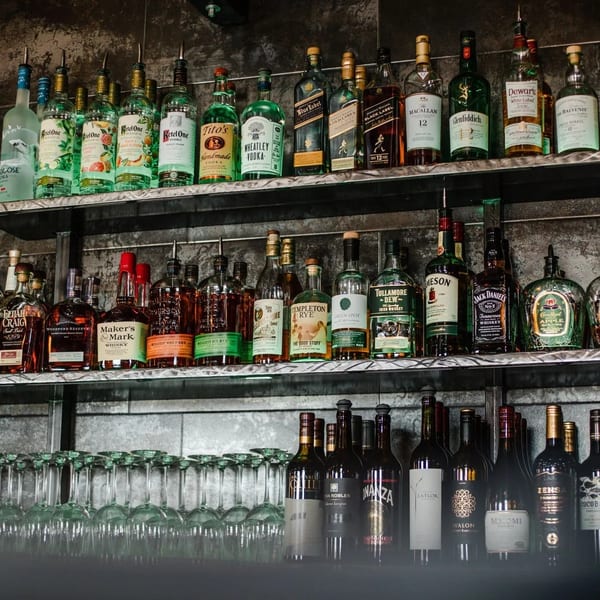 A well-stocked bar shelf with various liquor bottles, wine bottles, and rows of empty glasses.
