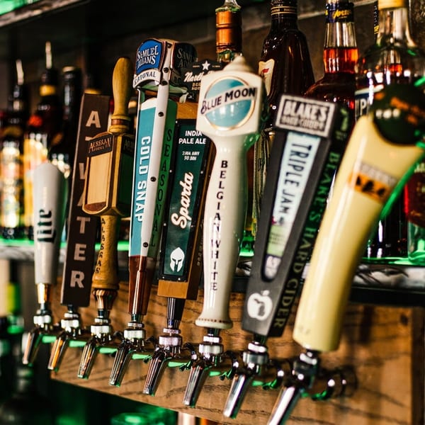 A close-up of several beer taps with various brand handles, and liquor bottles blurred in the background.